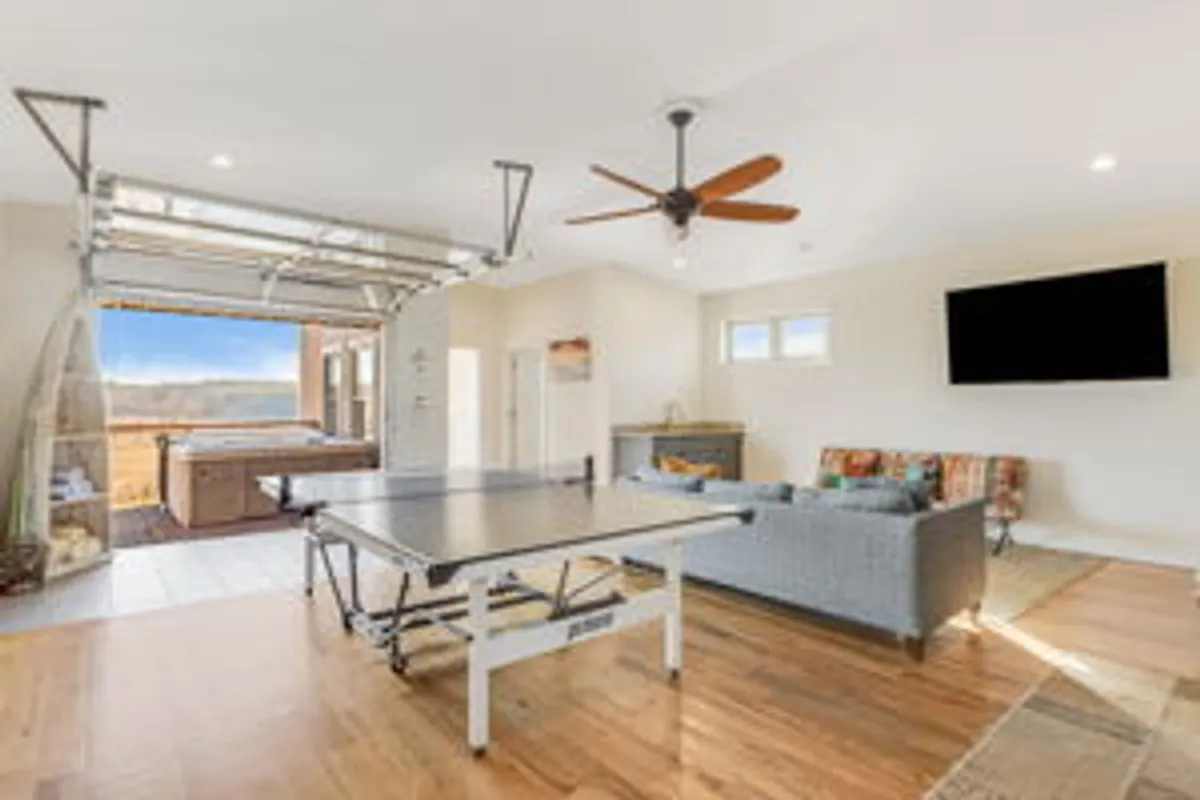 A vibrant recreational room featuring a ping pong table, a sofa area for relaxing, an open garage door revealing sunset views, and a hot tub. The space is casual and designed for group enjoyment and social activities.