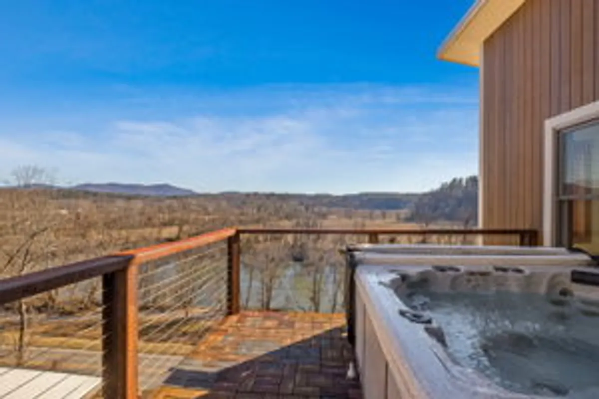 An exterior shot of a hot tub positioned on a wooden deck with unobstructed river views in the background. The setting exudes tranquility and the design encourages socializing or peaceful reflection in a scenic locale.