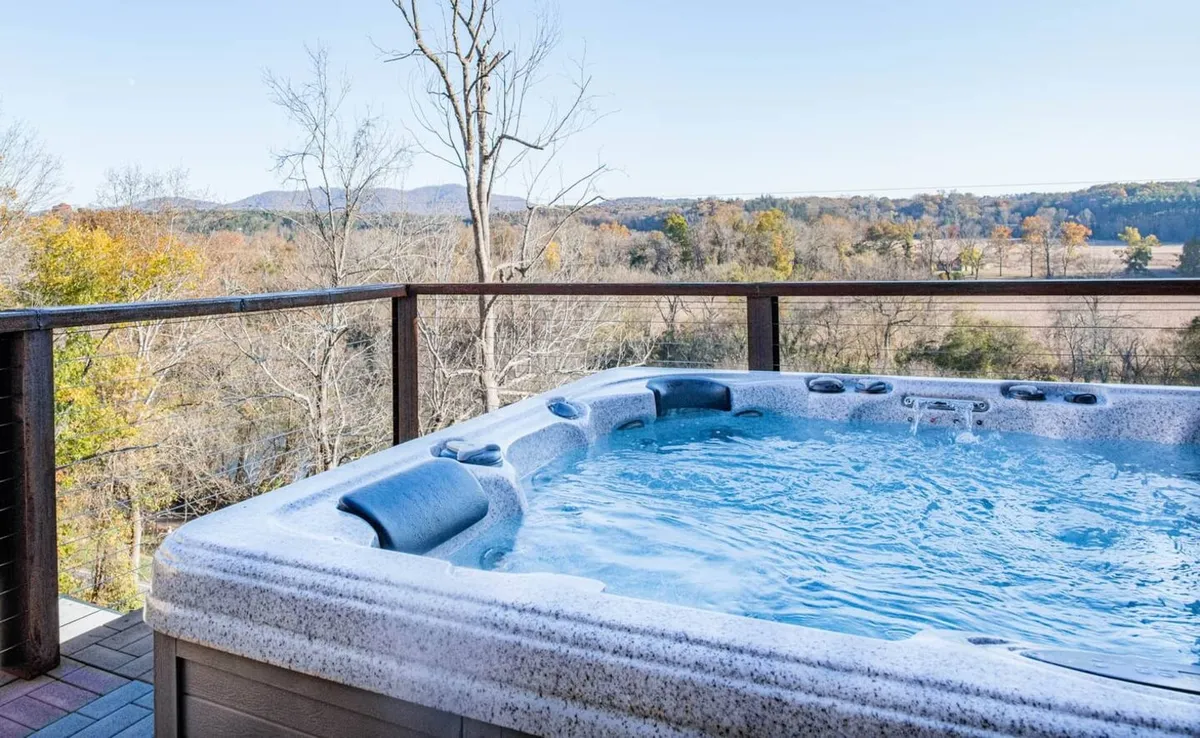 The image shows the exterior of a spacious single-family house nestled among lush trees, with a rooftop hot tub overlooking sweeping mountain vistas. The hot tub is positioned for stunning sunset and starlit views, inviting guests to relax and socialize while taking in the natural beauty of Asheville and the nearby Biltmore Estate.