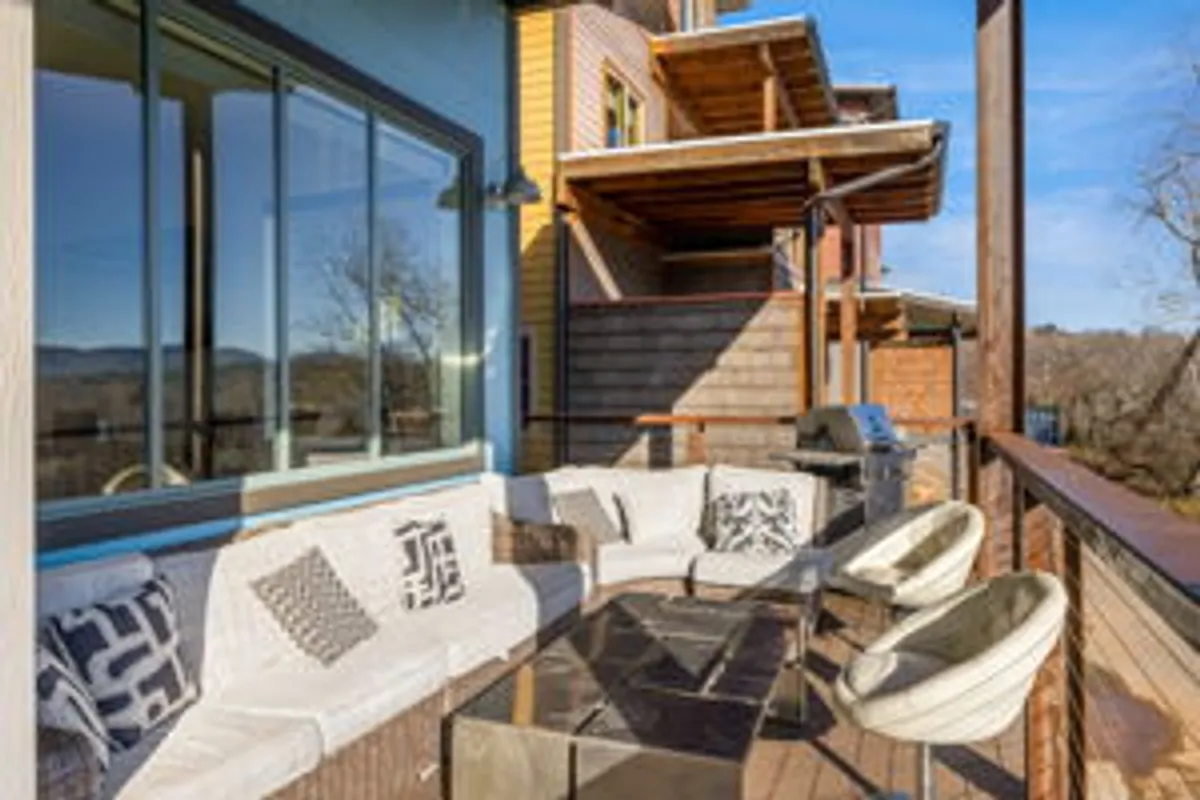 This image showcases a relaxed outdoor seating area with a glass table, surrounded by comfortable chairs. The patio is set against a scenic backdrop of woodland greenery and distant hills, bathed in gentle natural light. The setting is ideal for enjoying both morning coffee and evening gatherings, with clear sightlines to the landscape beyond.