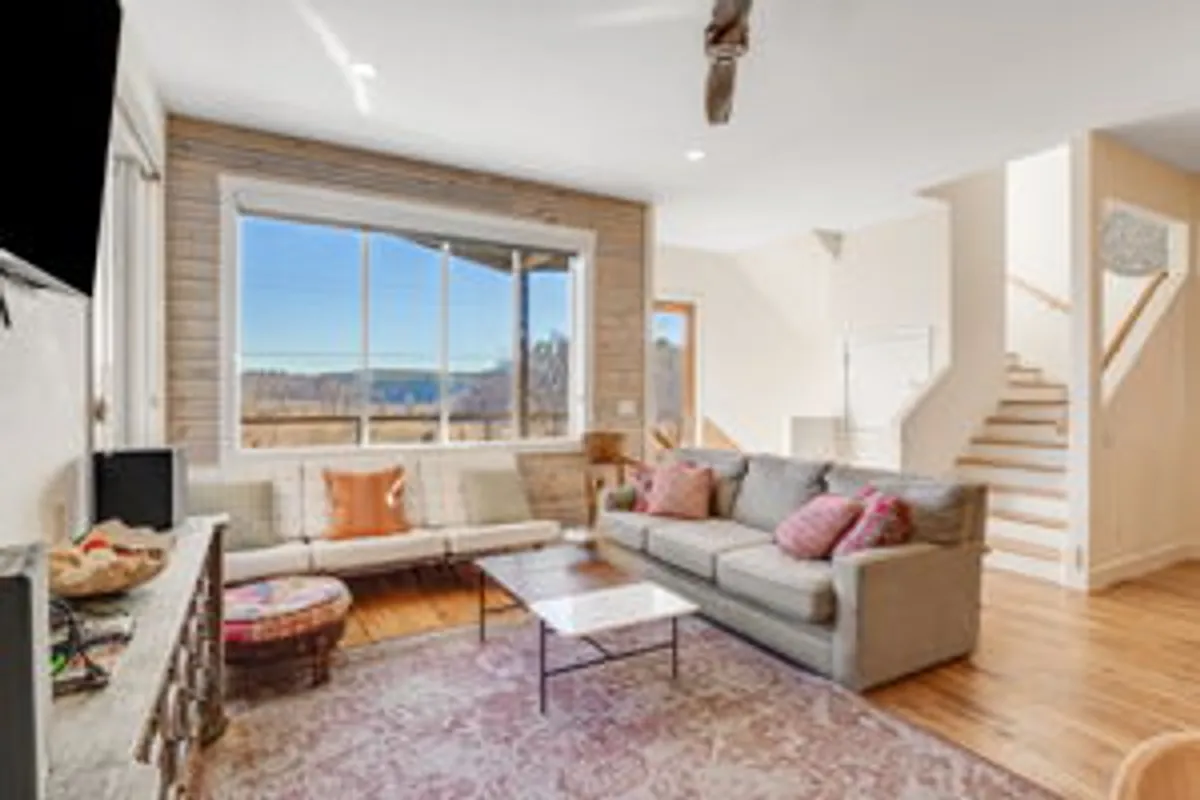 A welcoming living room is framed by a large window offering scenic views of the natural landscape. Comfortable seating, a coffee table, and a decorative rug create a serene setting for relaxation, while the window connects the interior space to mountain and woodland vistas.