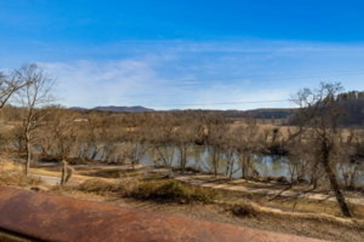 A wide view of the French Broad River framed by bare trees and distant mountain peaks. The tranquil river scene suggests opportunities for hiking, wildlife watching, and sunset viewing, just steps from the home.