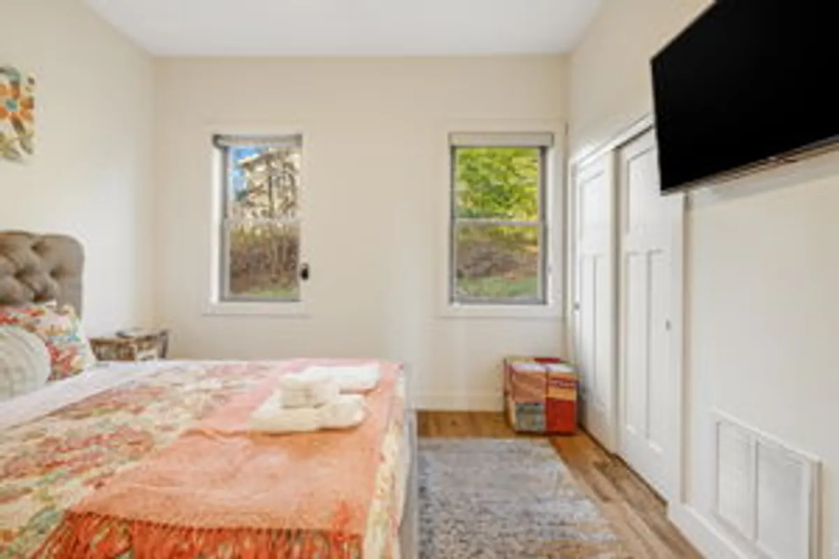 A bright bedroom featuring a bed with colorful bedding, large windows letting in natural light, and a wall-mounted television. The atmosphere is tranquil, with a playful use of color and soft textures, echoing the inviting and comfortable theme of the section.