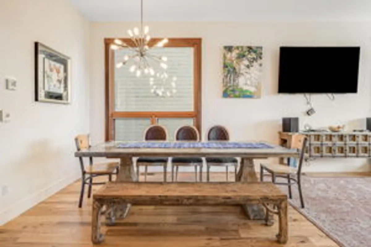 A welcoming dining area centered around a wooden table with a chandelier overhead, surrounded by art and a television. The room exudes warmth and is set up for both family meals and festive gatherings.
