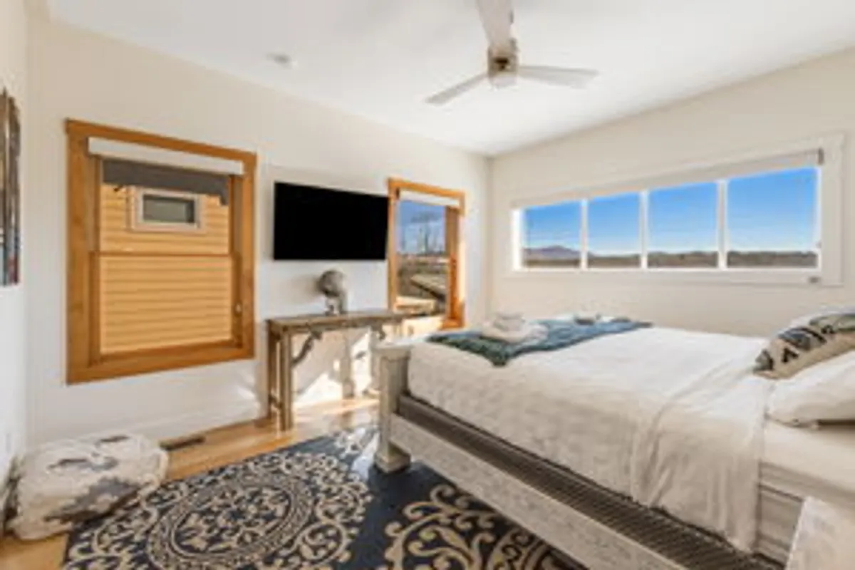 A comfortable bedroom with a large window framing a beautiful landscape, inviting in natural light. The room includes a bed, TV, wooden table, and artwork, with a peaceful, cozy ambiance ideal for relaxation and taking in the sunset or mountain views.