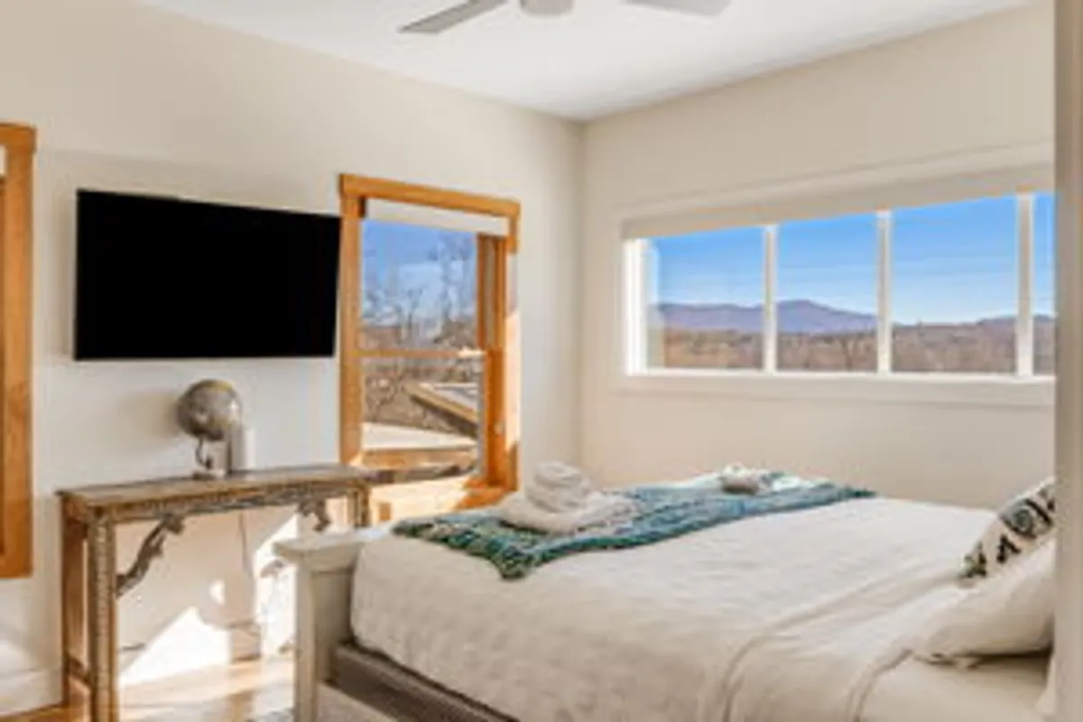 This image features a serene bedroom with a large window revealing an impressive mountain view, a neatly arranged bed, a decorative table, and a TV. The window immerses the interior in natural light and connects the space directly to the stunning landscape outside.