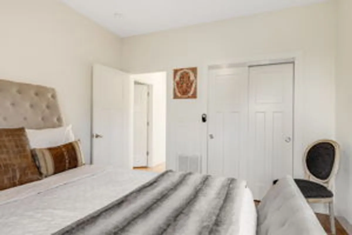 A serene bedroom with a plush headboard, decorative pillows, wall art, and a cozy chair in the corner. Doorways lead to other areas, adding a sense of spaciousness and privacy, while the decor feels both calming and inviting.