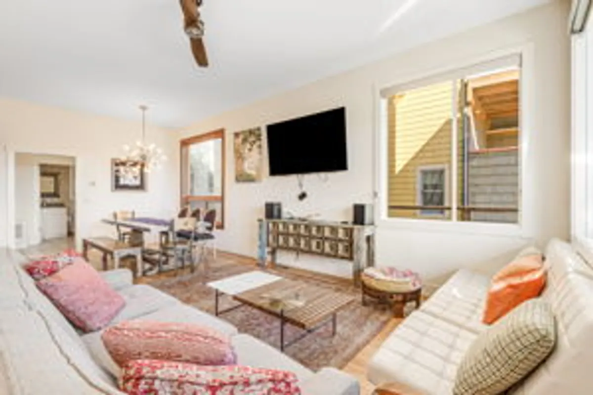 A cozy, open-concept living and dining area is revealed, with a plush sofa adorned with colorful cushions, a stylish dining table, a TV, and a striking chandelier overhead. The layout encourages relaxation, conversation, and shared meals, perfectly reflecting the communal atmosphere described in the section.